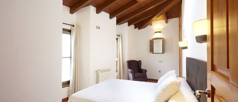 Un dormitorio con una cama grande, una silla, una mesita de noche con un libro, una ventana con cortinas y techo de madera.