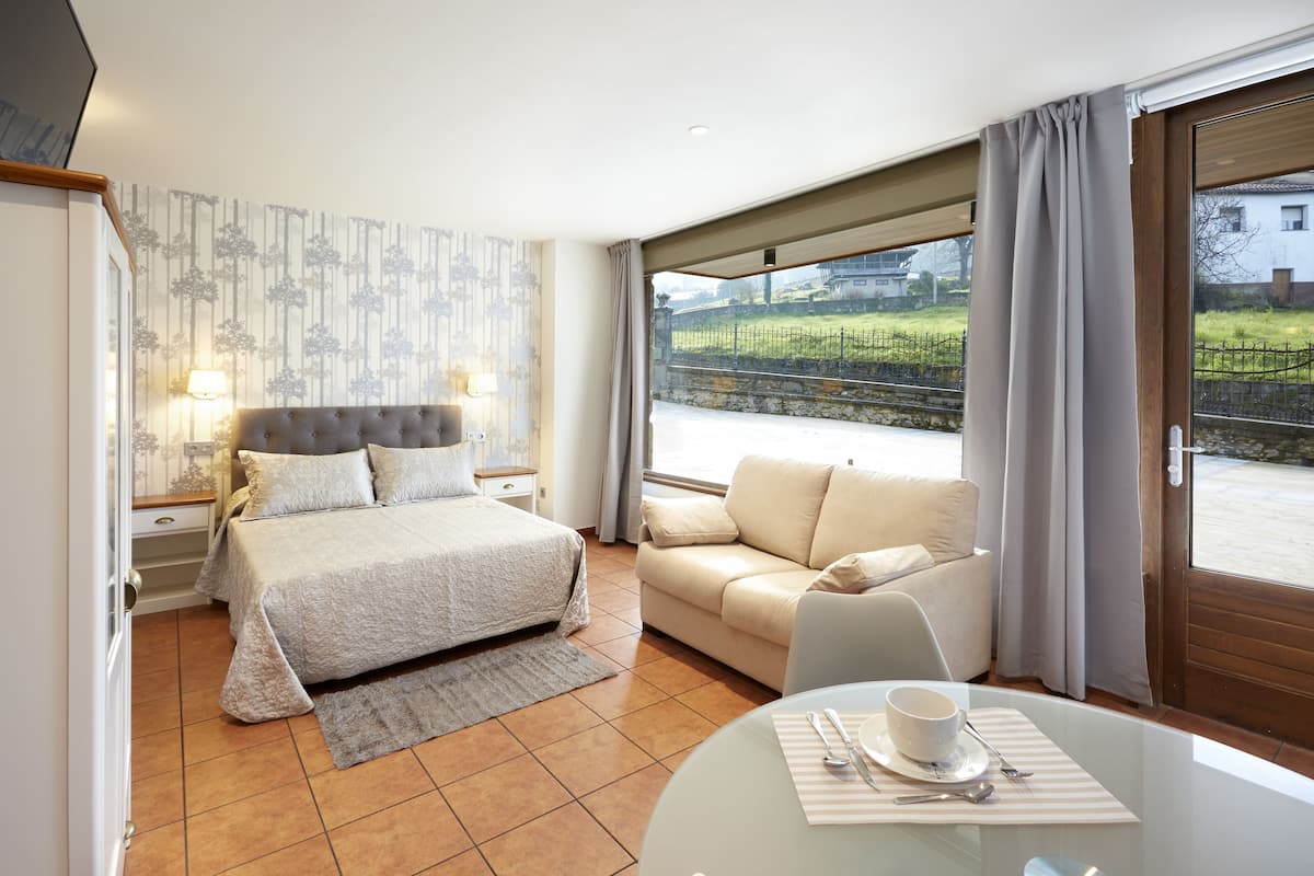 A hotel room with a bed, a sofa, a dining table with cutlery, and a view of a garden and buildings through large windows.