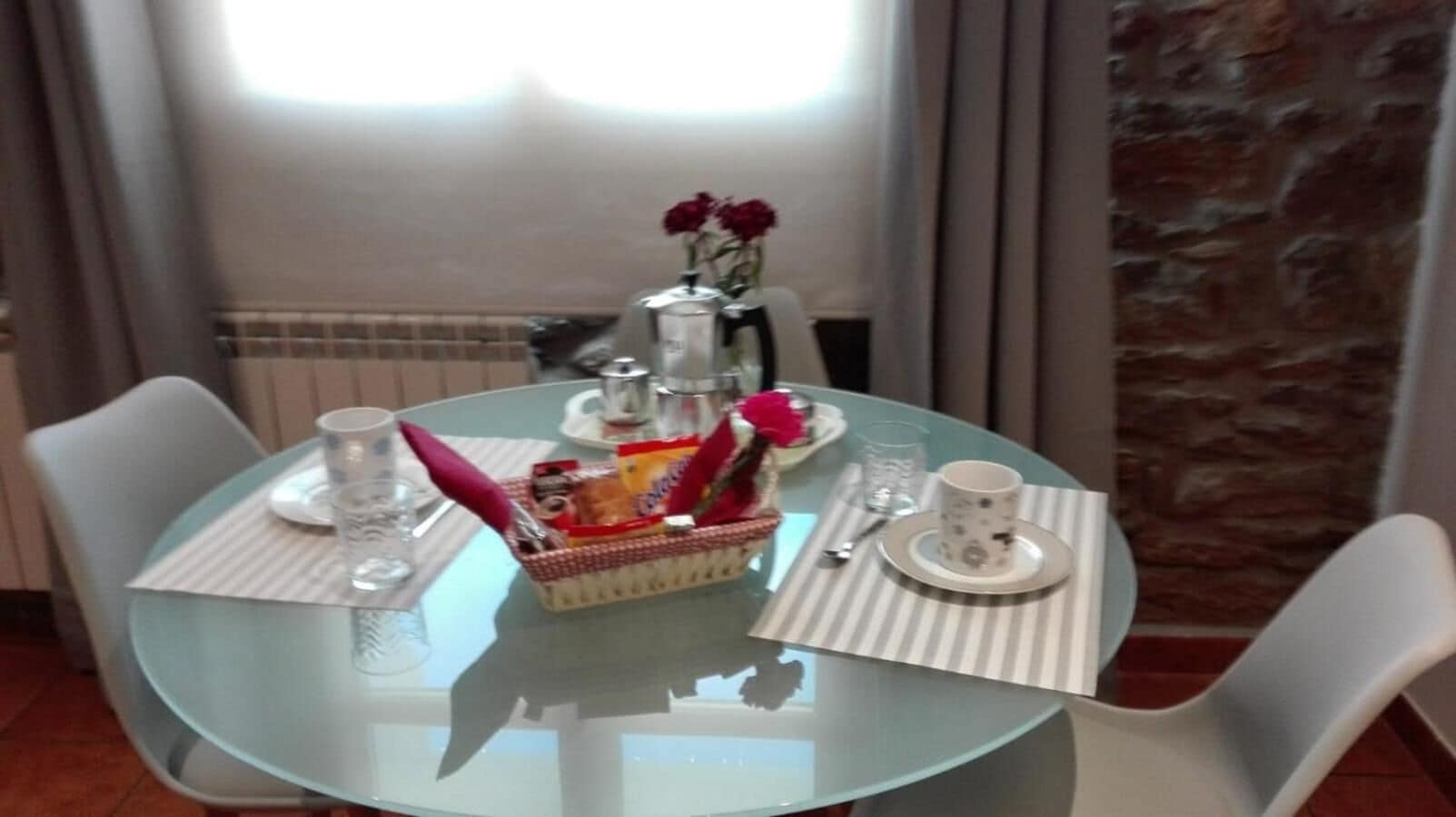 Une table ronde en verre, des chaises blanches, un panier de collations, un vase rempli de fleurs et une cafetière.