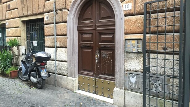 A stone building with a large, ornate brown door and a metal gate. A scooter is parked in front. - Riari Trastevere Apartment (Rome)