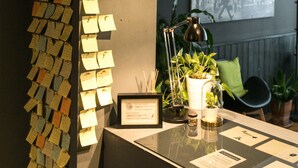 A reception desk with a wall of notes, a framed certificate, a potted plant, and a chair.