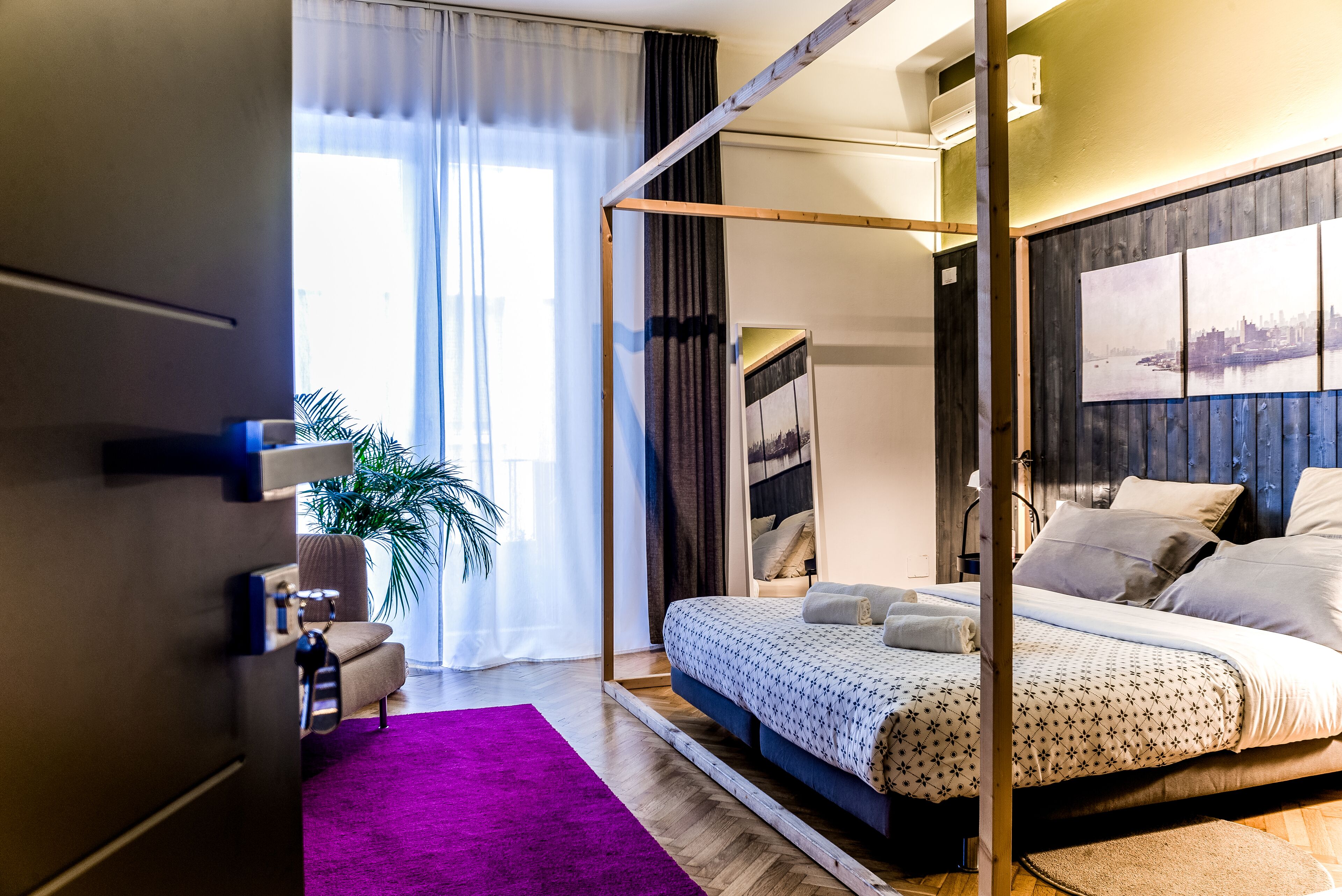 Une chambre d’hôtel moderne avec un lit à baldaquin, une grande fenêtre avec des rideaux, un tapis violet et un tableau représentant un paysage urbain accroché au mur.