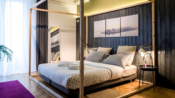 A modern bedroom with a canopy bed, a cityscape artwork on the wall, and a small bedside table with a lamp.