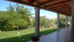 Un patio couvert doté d’un sol carrelé, de colonnes en pierre et offrant une vue sur un jardin aménagé avec des arbres et des cactus.