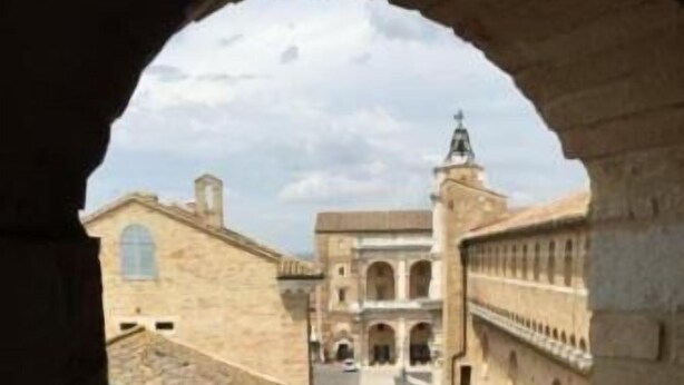 A narrow alleyway with stone buildings, a visible archway, and a distant view of a larger building with a tower. - Pensione Piemonte (Loreto)