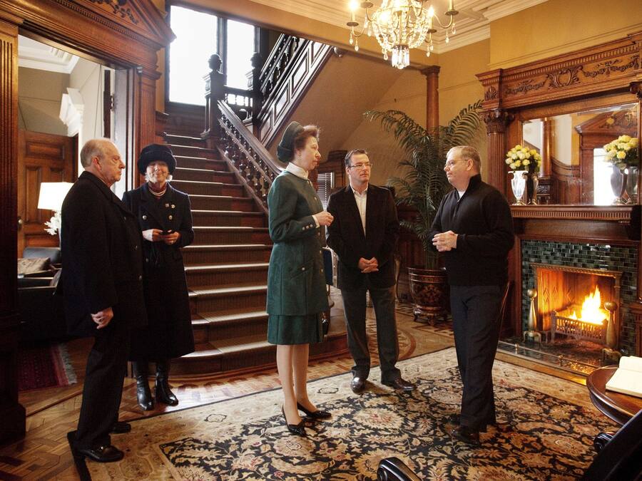 Un grupo de personas en un ambiente formal, con una escalera, una chimenea y un candelabro.