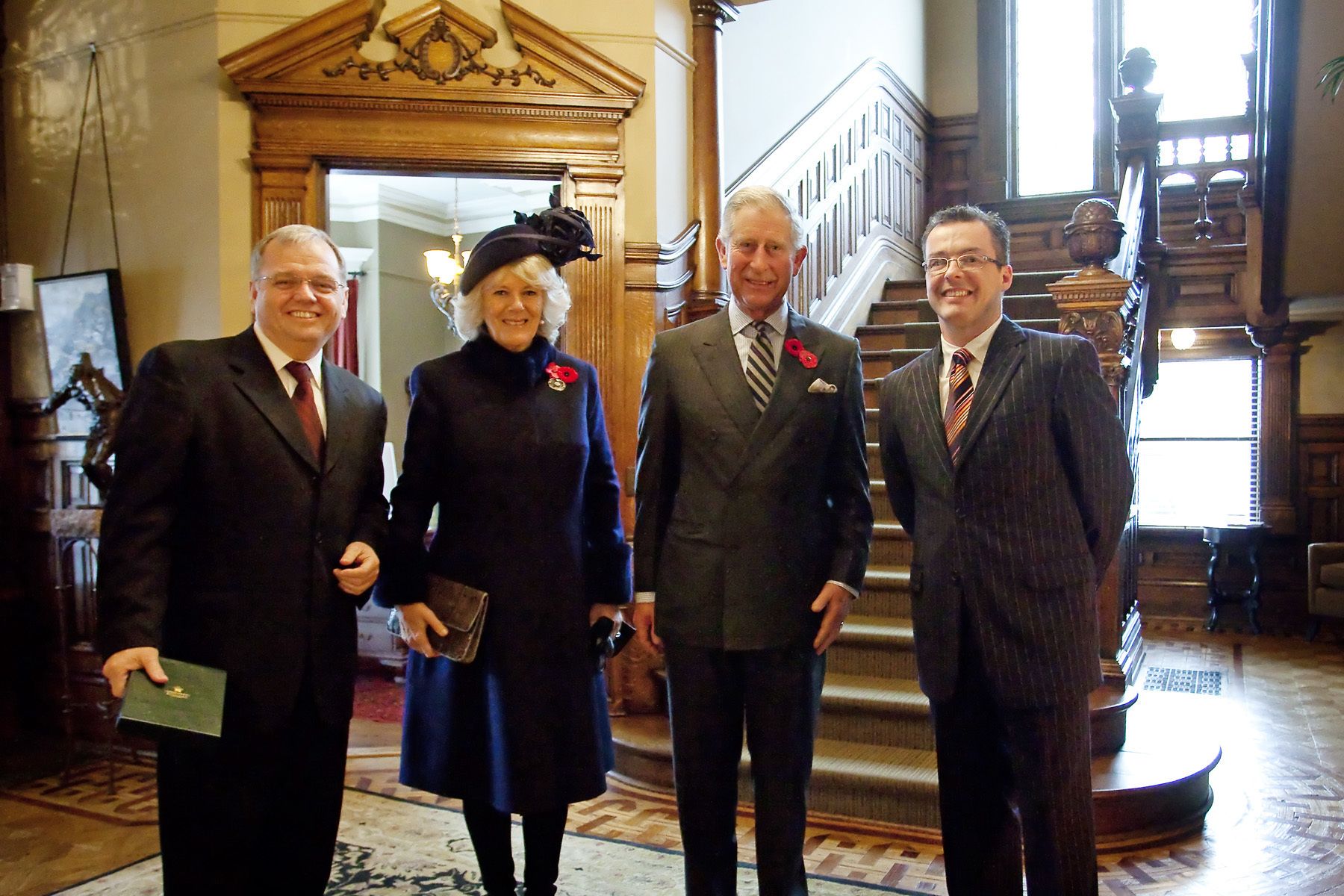 A group of four people in a formal setting with a grand staircase and ornate woodwork.