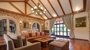 Un salón amplio con pared de piedra, vigas de madera, una araña y ventanas grandes con cortinas.