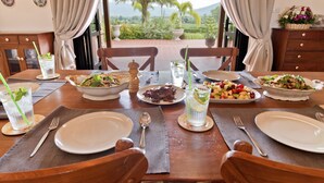 Una mesa de comedor dispuesta con platos, cubiertos y bebidas, con vistas a un paisaje pintoresco con palmeras y montañas.