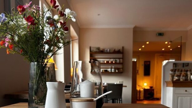 A dining table with a white pitcher, a white cup, and a white bowl on a saucer. A vase with flowers is on the table. - Hotel Finkenhof - Haus Meersmannufer (Hannover)