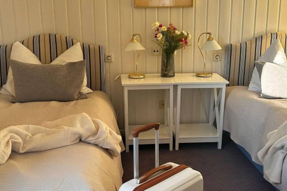 A hotel room with two beds, a small table with a vase of flowers, two lamps, and a suitcase.