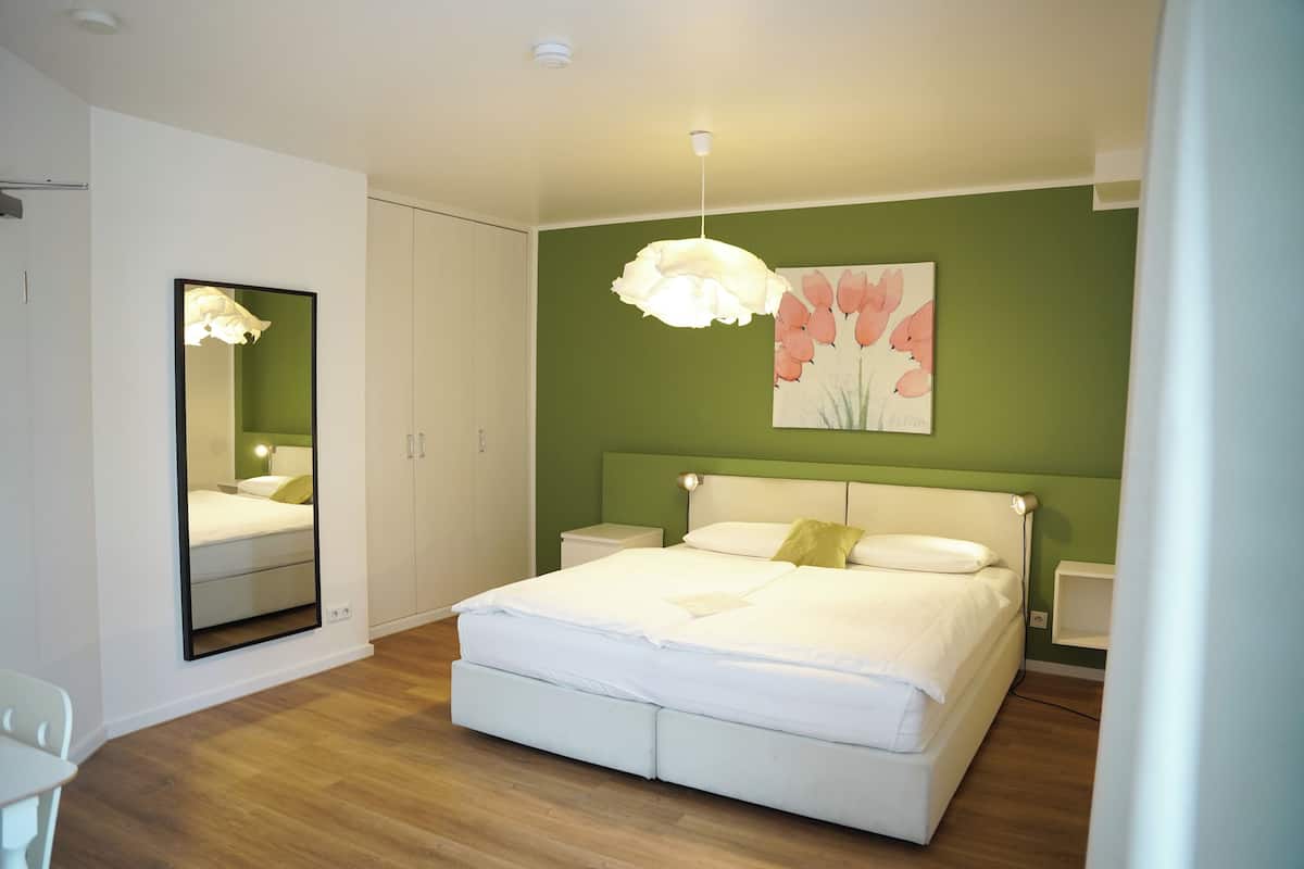 A bedroom with a large bed, a bedside table, a nightstand, a mirror, and a wall-mounted light fixture.