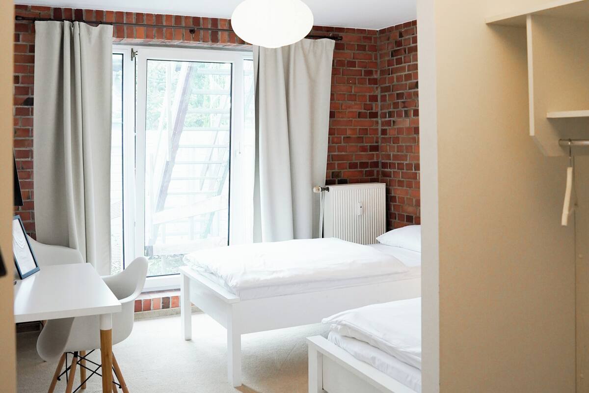 A room with a white desk, two beds, a radiator, and a window with curtains.