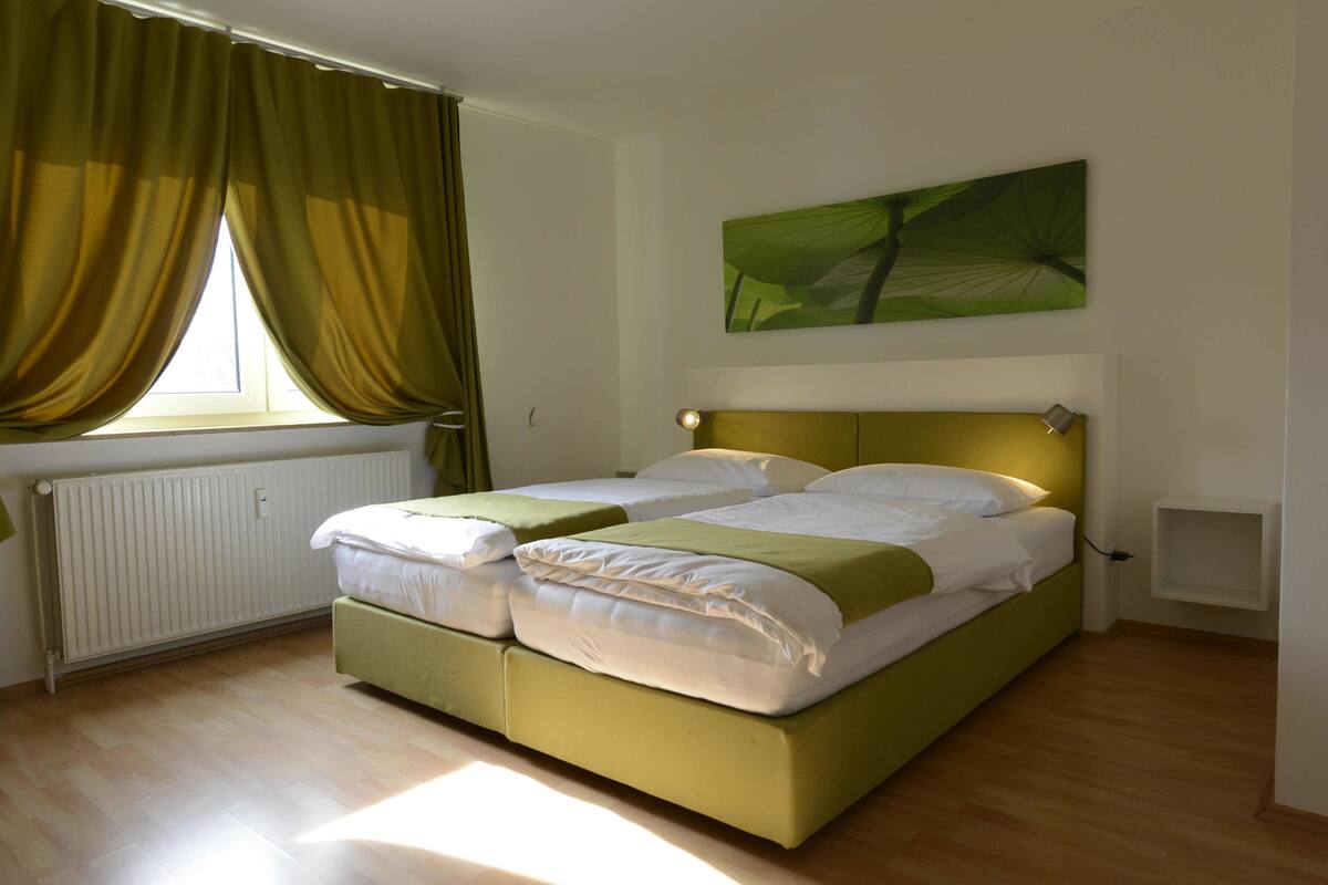 A double bed with a green headboard in a room with wooden flooring, a window with curtains, and a wall-mounted light fixture.