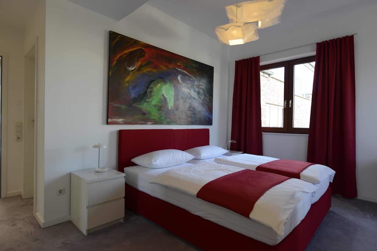 A double bed with red headboard and white bedding, a nightstand with a lamp, a large abstract painting on the wall, and a window with red curtains.