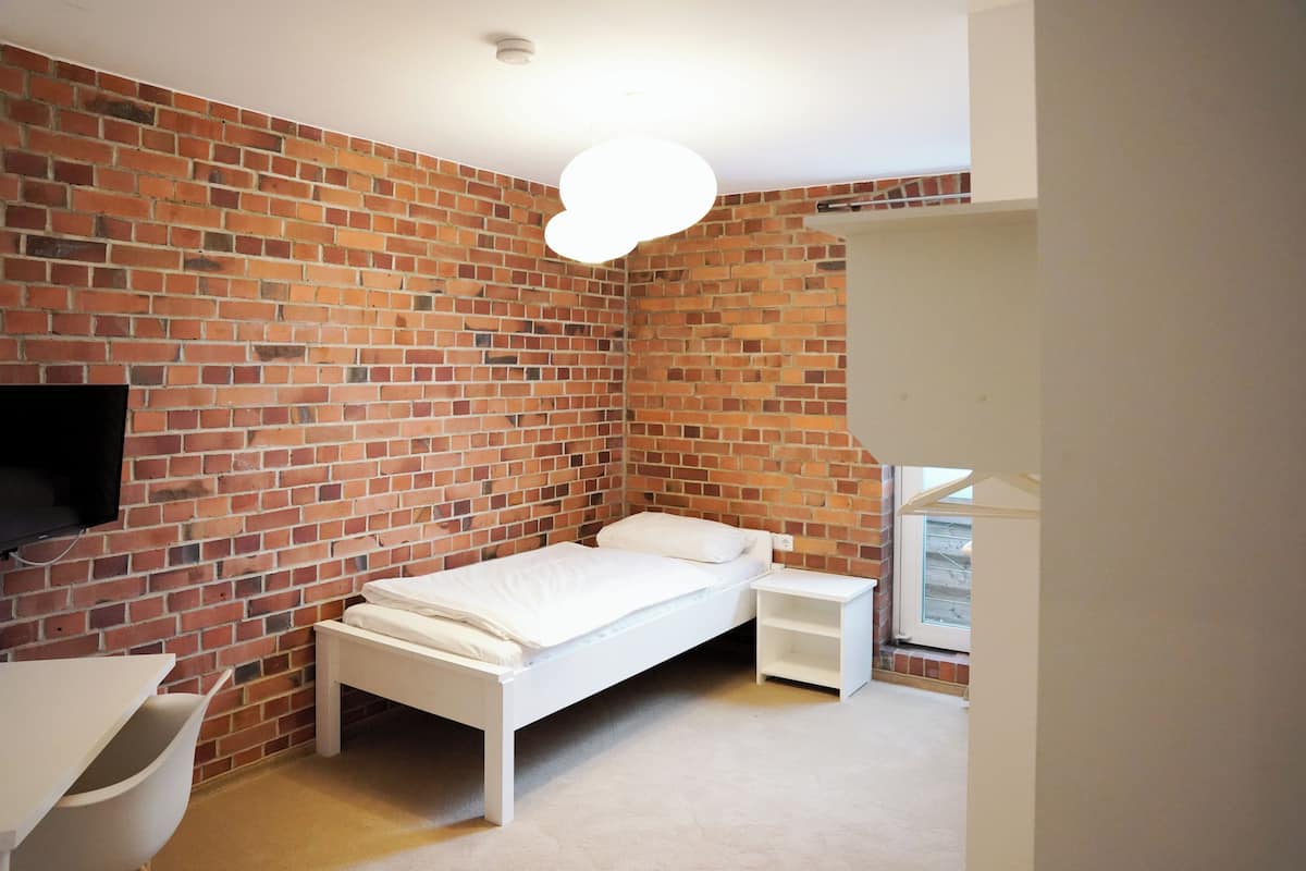 A bedroom with a brick wall, a white bed, a bedside table, a TV mounted on the wall, and a window.