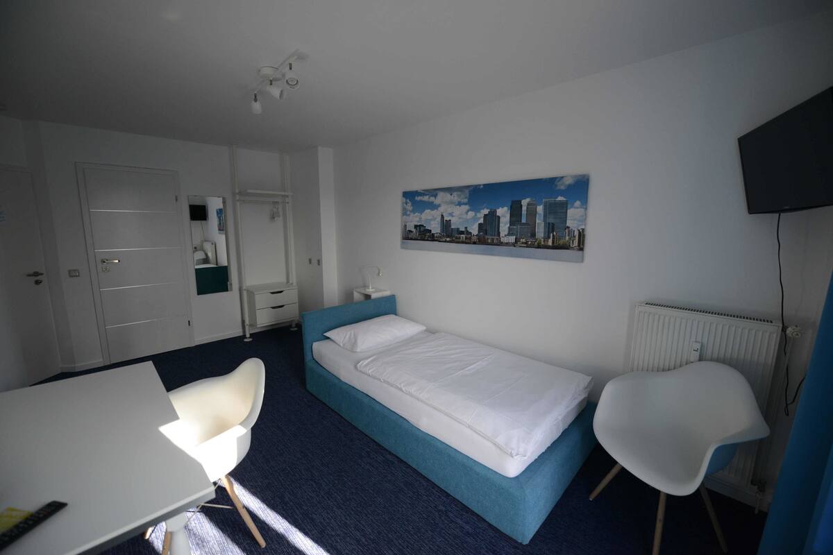 A hotel room with a single bed, a white desk, a chair, a radiator, and a cityscape painting on the wall.