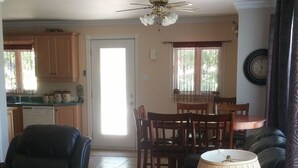 Un salon avec un canapé en cuir noir, une table basse en bois et un ventilateur de plafond. On aperçoit à l’arrière-plan un coin repas avec des chaises et une table en bois.