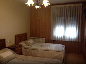 Una habitación con dos camas, cabecera de madera, mesita de noche, radiador y ventana con cortinas.