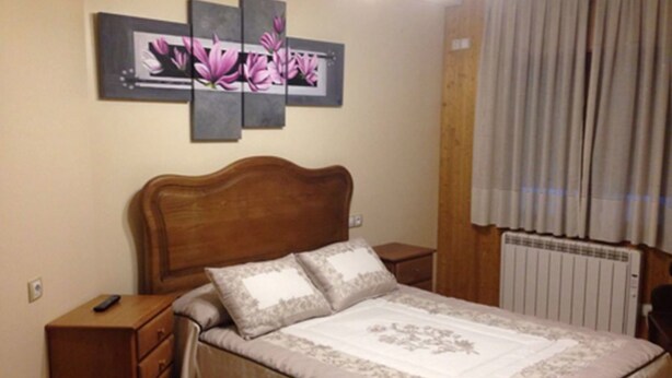 Een slaapkamer met een houten hoofdbord, een bed met bloemenbeddengoed, een nachtkastje met een afstandsbediening en een radiator.