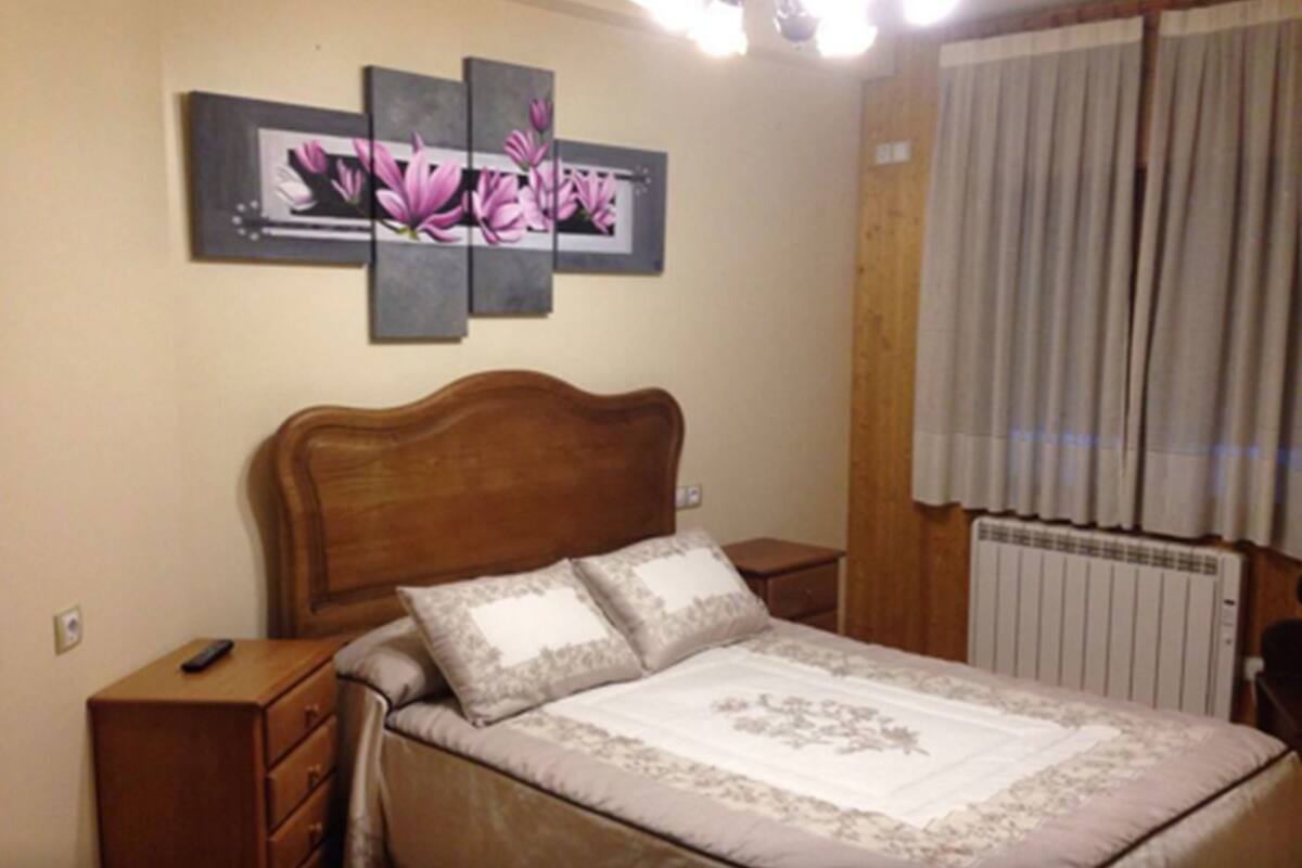 Une chambre avec une tête de lit en bois, un lit recouvert d’une literie à motifs floraux, une table de chevet avec une télécommande et un radiateur.