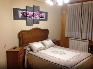 Quarto com cabeceira de madeira, cama com roupa de cama florida, mesa de cabeceira com controle remoto e radiador.