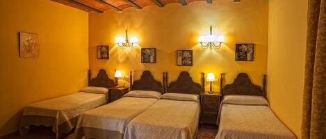 A room with four single beds, each with a wooden headboard and a bedside table with a lamp. The room has a tiled floor and a wooden ceiling with exposed beams.