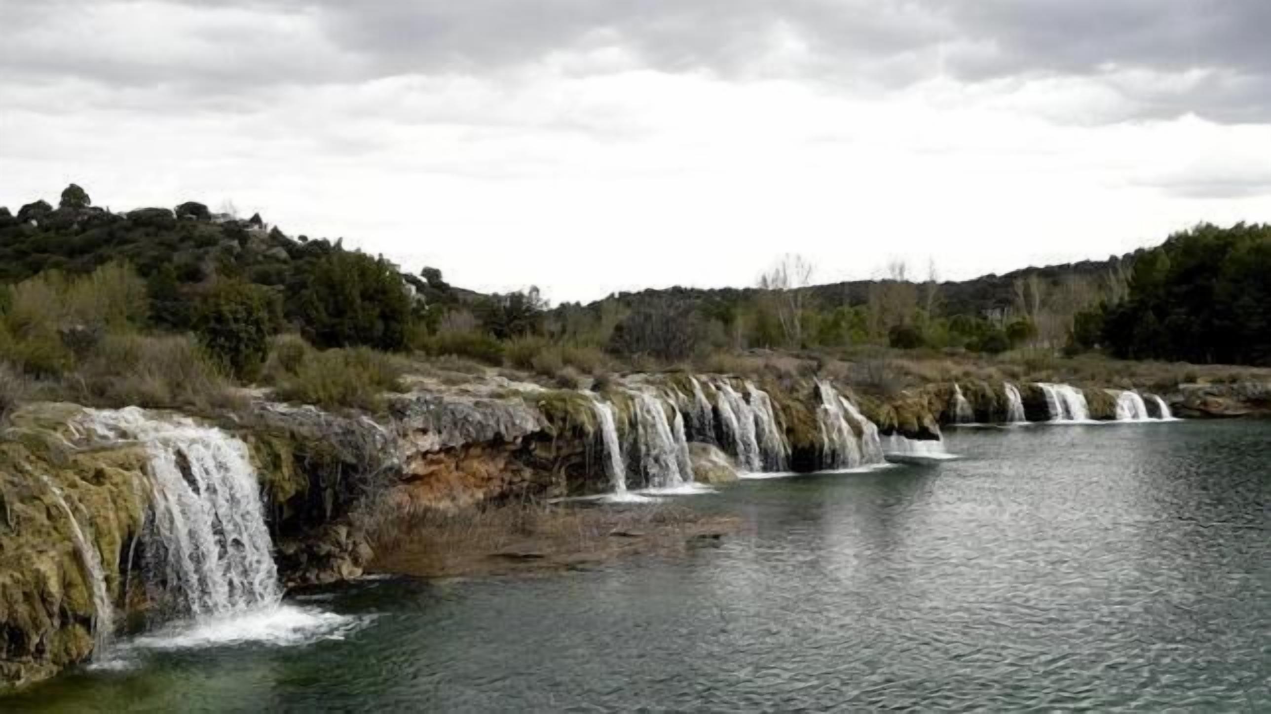 Una serie de cascadas que desembocan en un río tranquilo, rodeado de terreno rocoso y vegetación escasa.