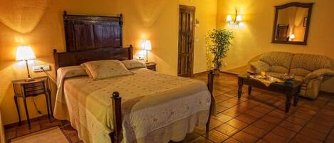 A cozy bedroom with a large bed, bedside lamps, a wooden headboard, a small table with a plant, and a sofa.