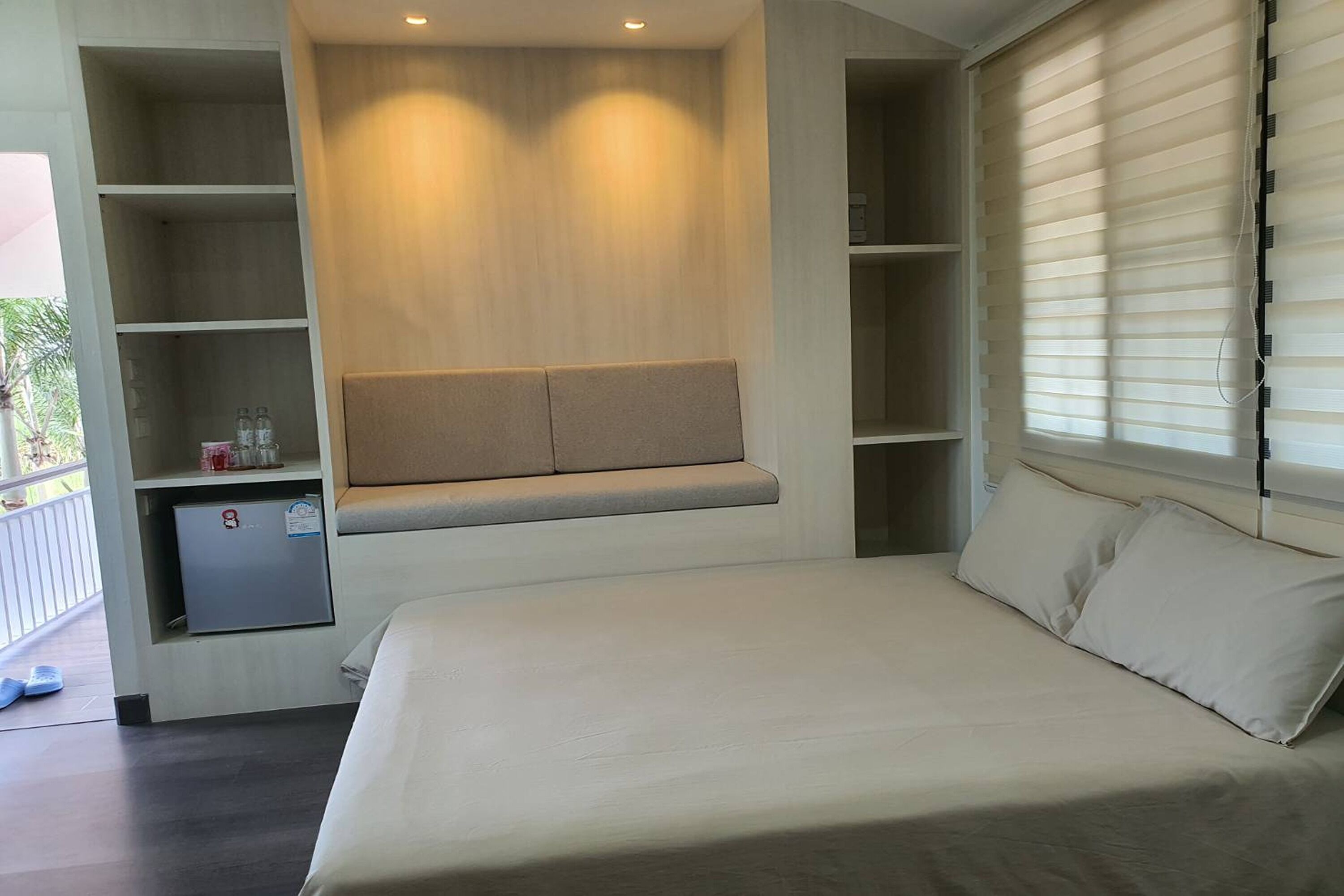 Un dormitorio con cama, estanterías empotradas, un refrigerador pequeño y una ventana con persianas.