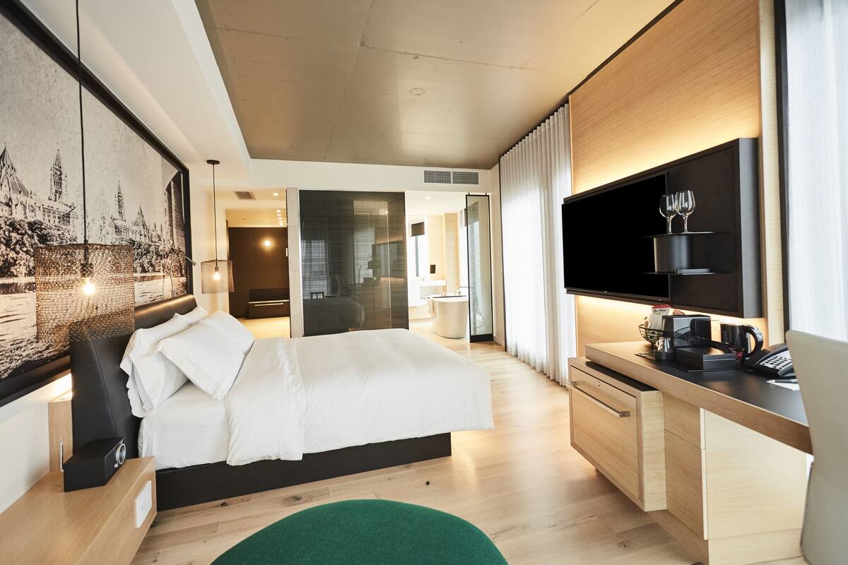 A modern hotel room with a large bed, a desk with a flat-screen TV, and a cityscape artwork on the wall.