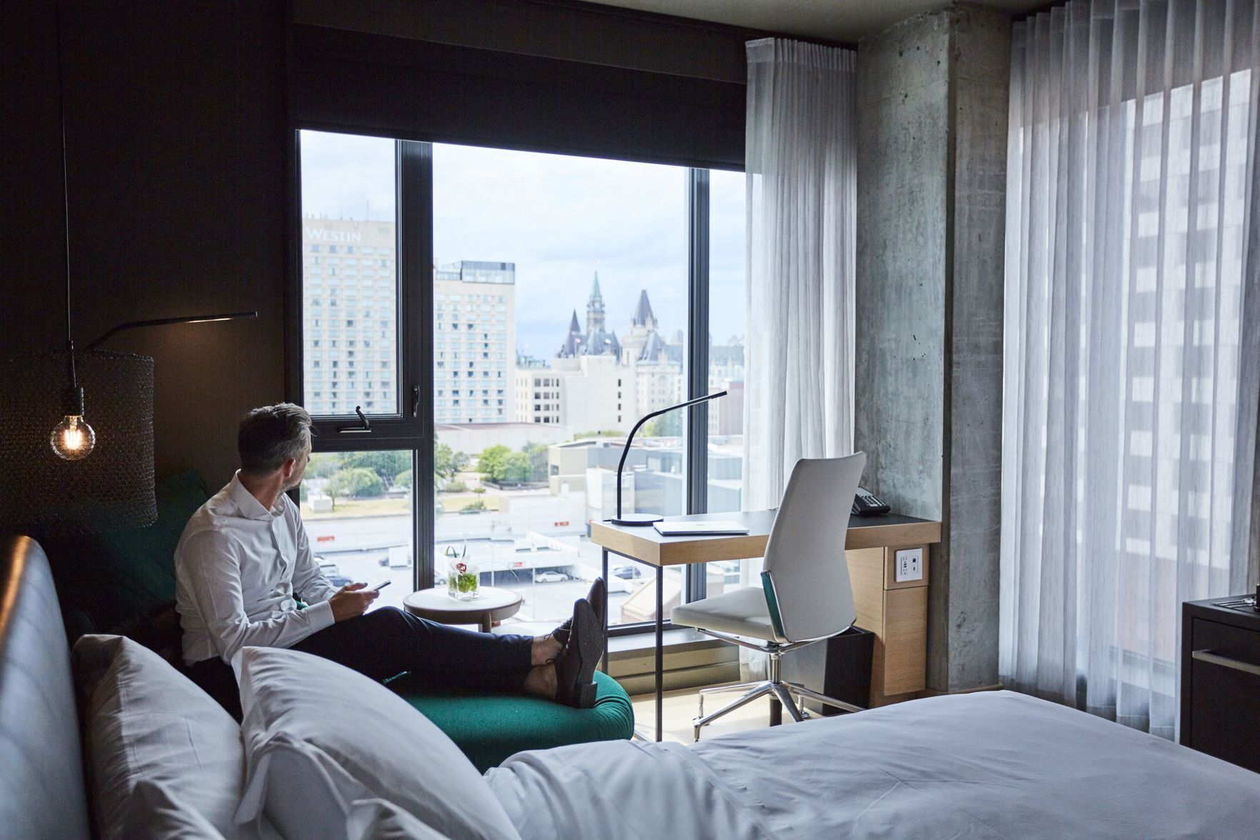 Habitación de hotel con cama, escritorio, silla, lámpara y vista a la ciudad.