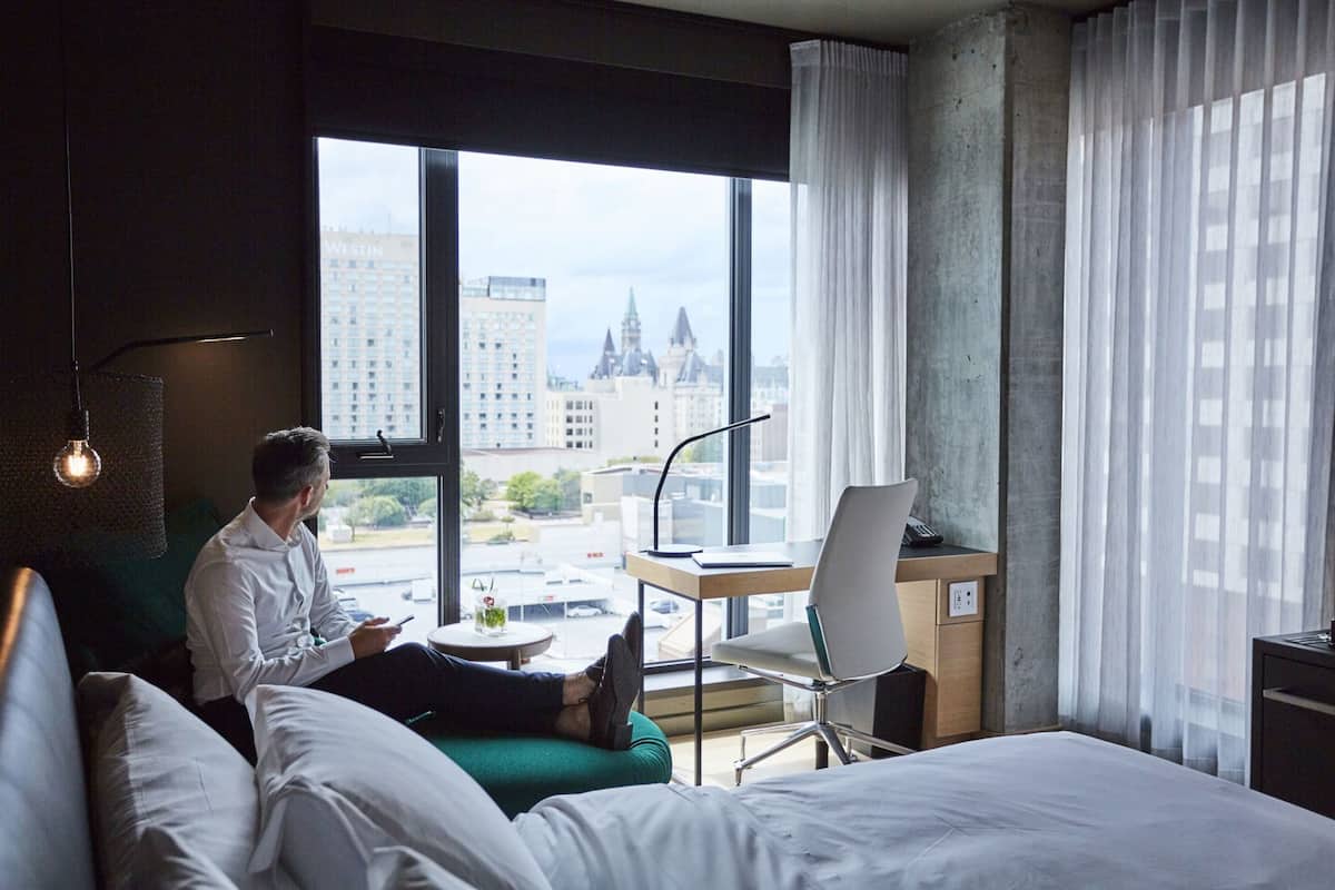 Quarto de hotel com cama, escrivaninha com cadeira, luminária e vista para a paisagem urbana.