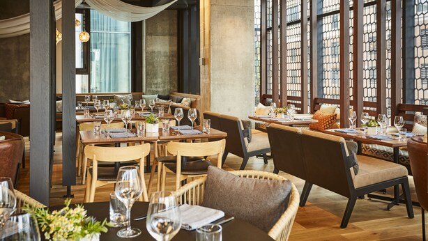 Un restaurante con mesas y sillas de madera, ventanas amplias con vistas y una lámpara colgante.