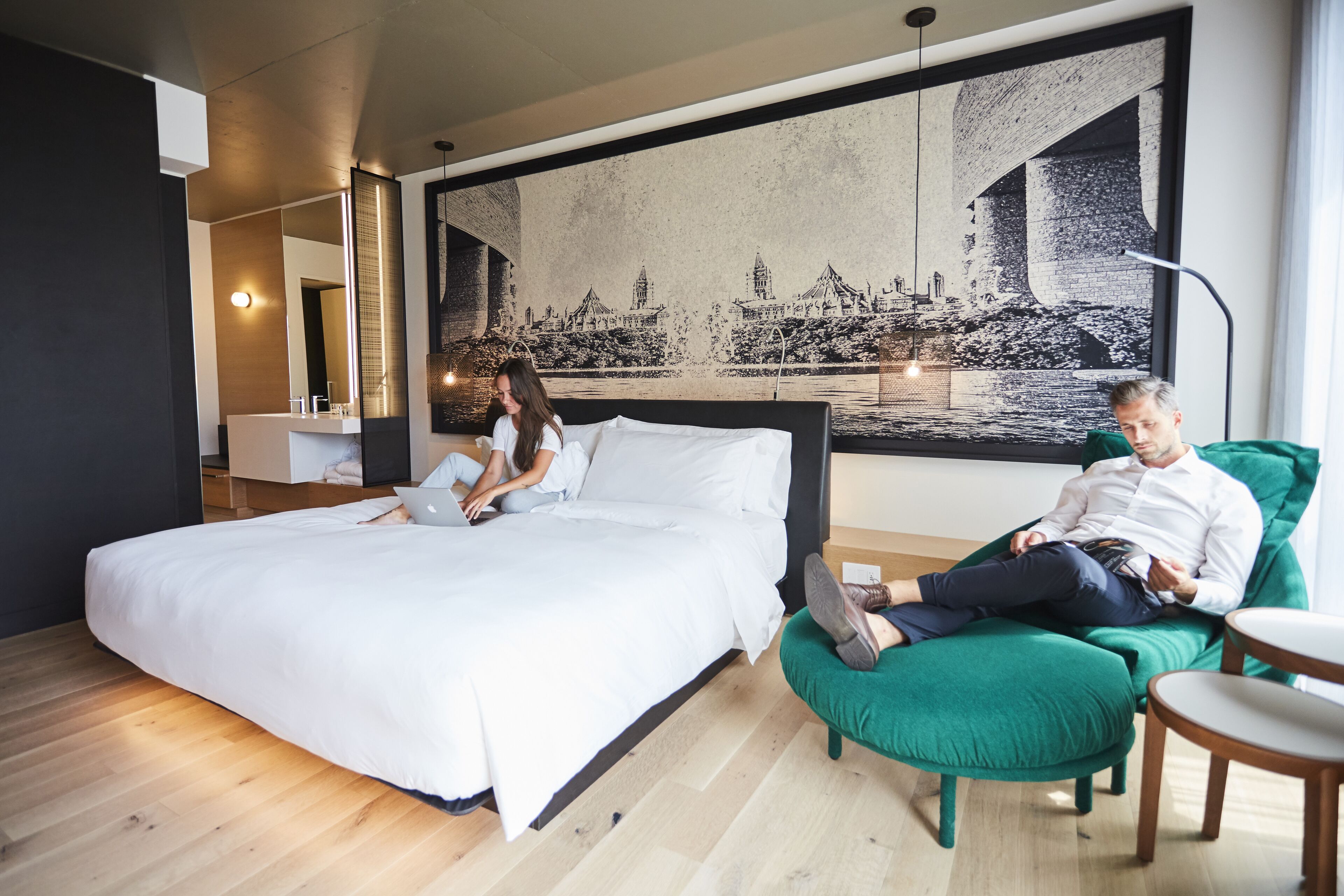A modern hotel room with a large bed, a green armchair, and a cityscape mural on the wall.