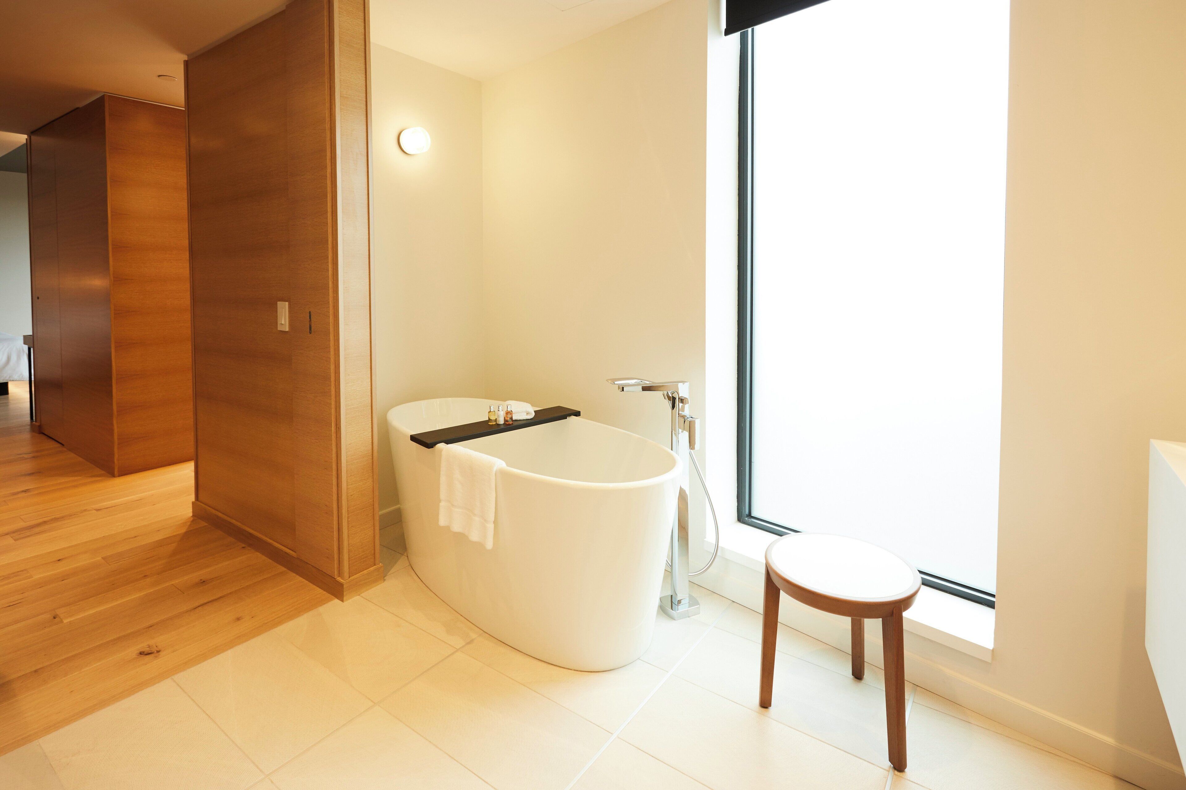 A modern bathroom with a freestanding bathtub, a small wooden stool, and a large window providing natural light.