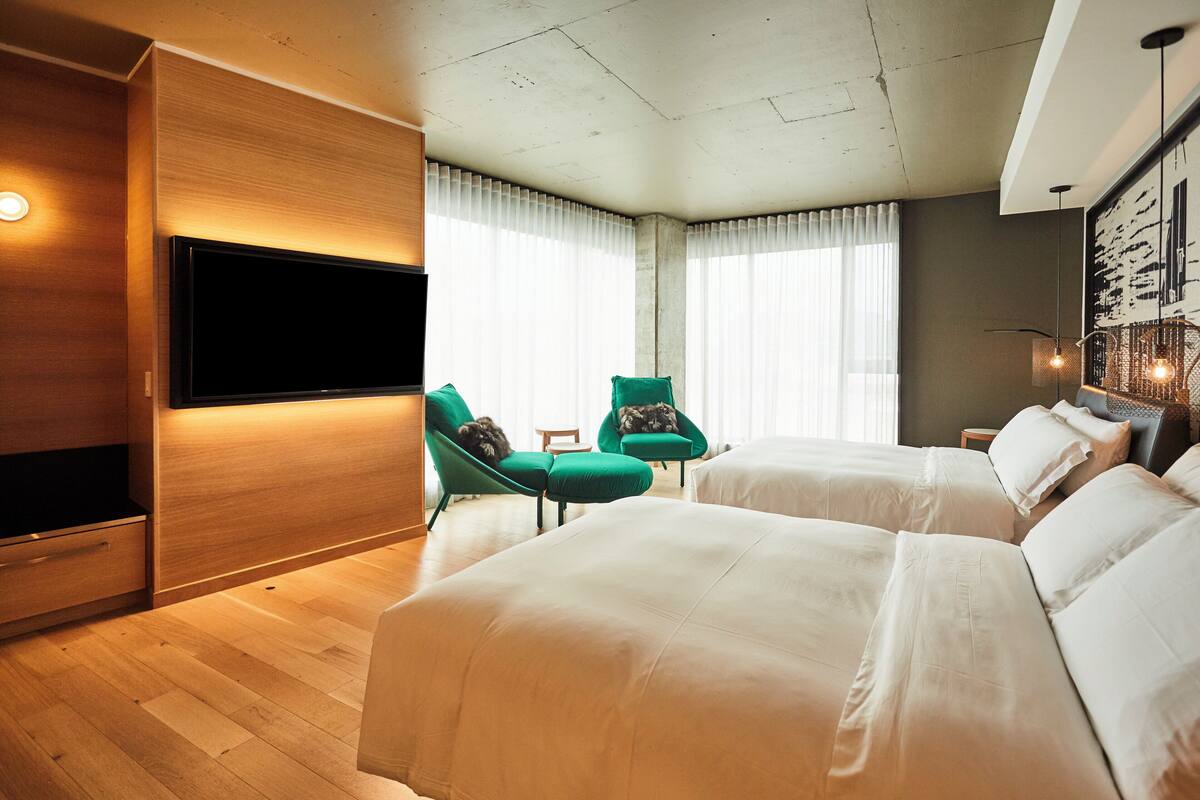 A hotel room with two beds, a flat-screen TV, green armchairs, and a large window with curtains.