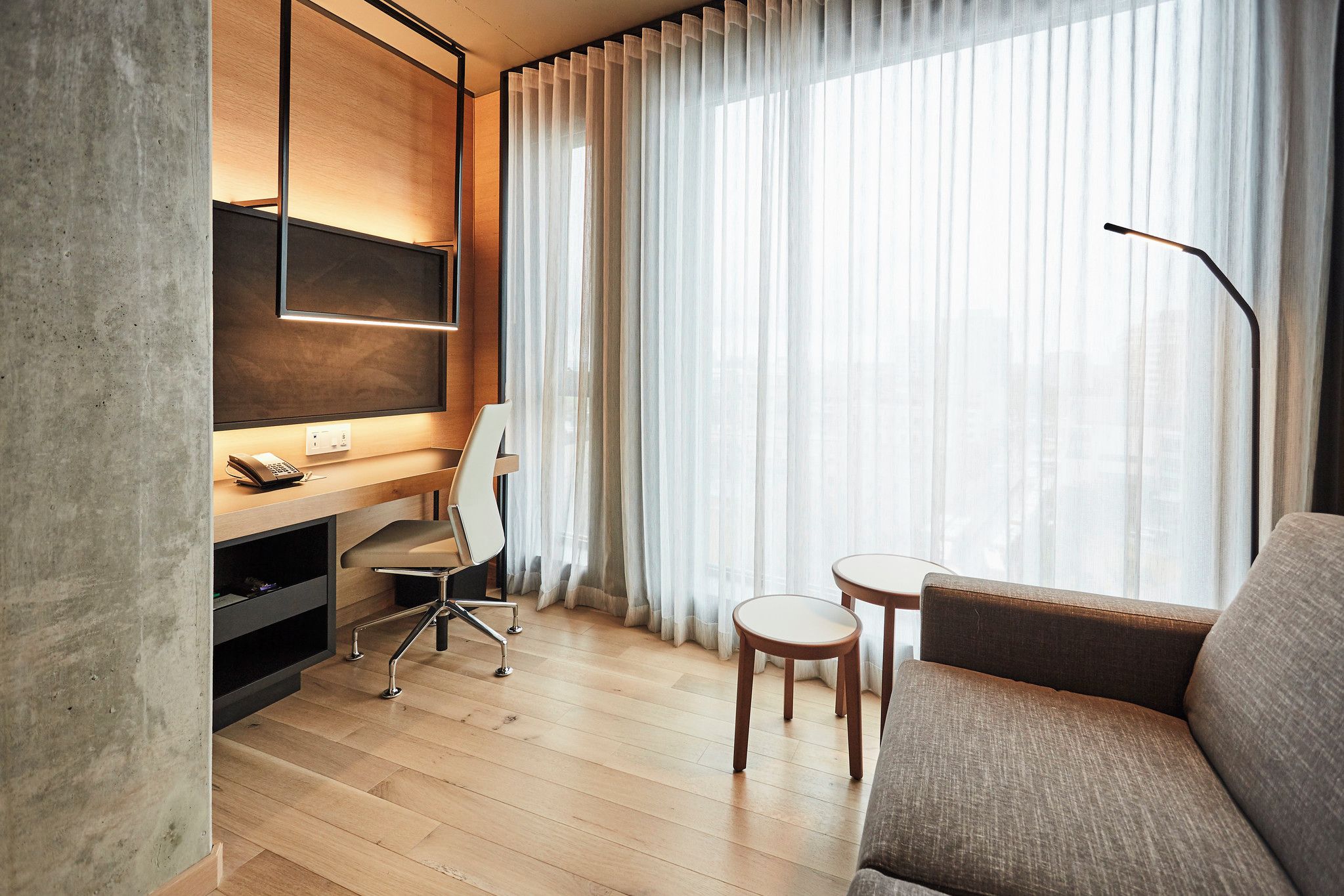 Habitación de hotel moderna con escritorio de madera, una silla, un sofá y un televisor fijado a la pared.
