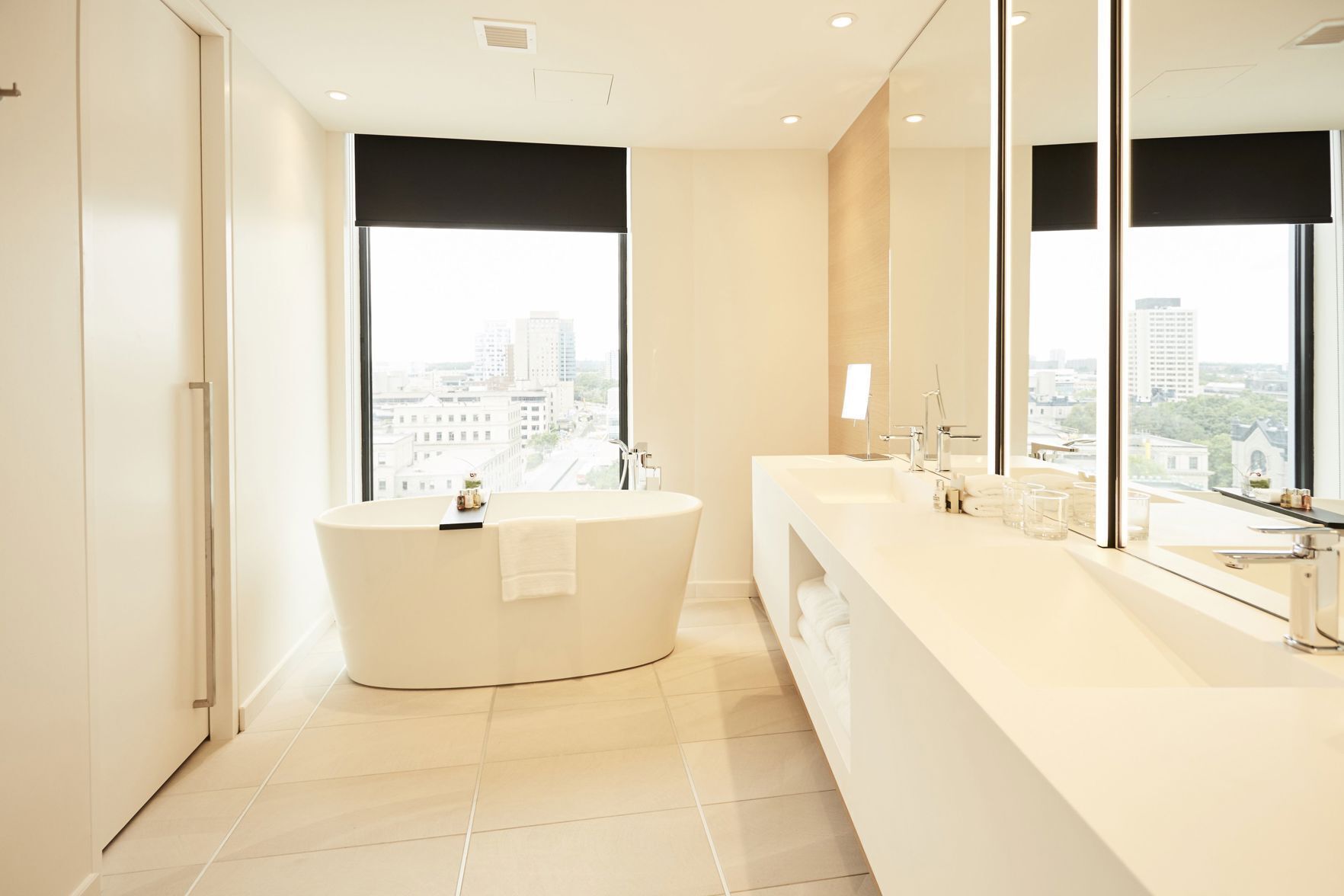 Un baño moderno con una bañera independiente, un espejo grande y vistas a la ciudad a través de las ventanas.