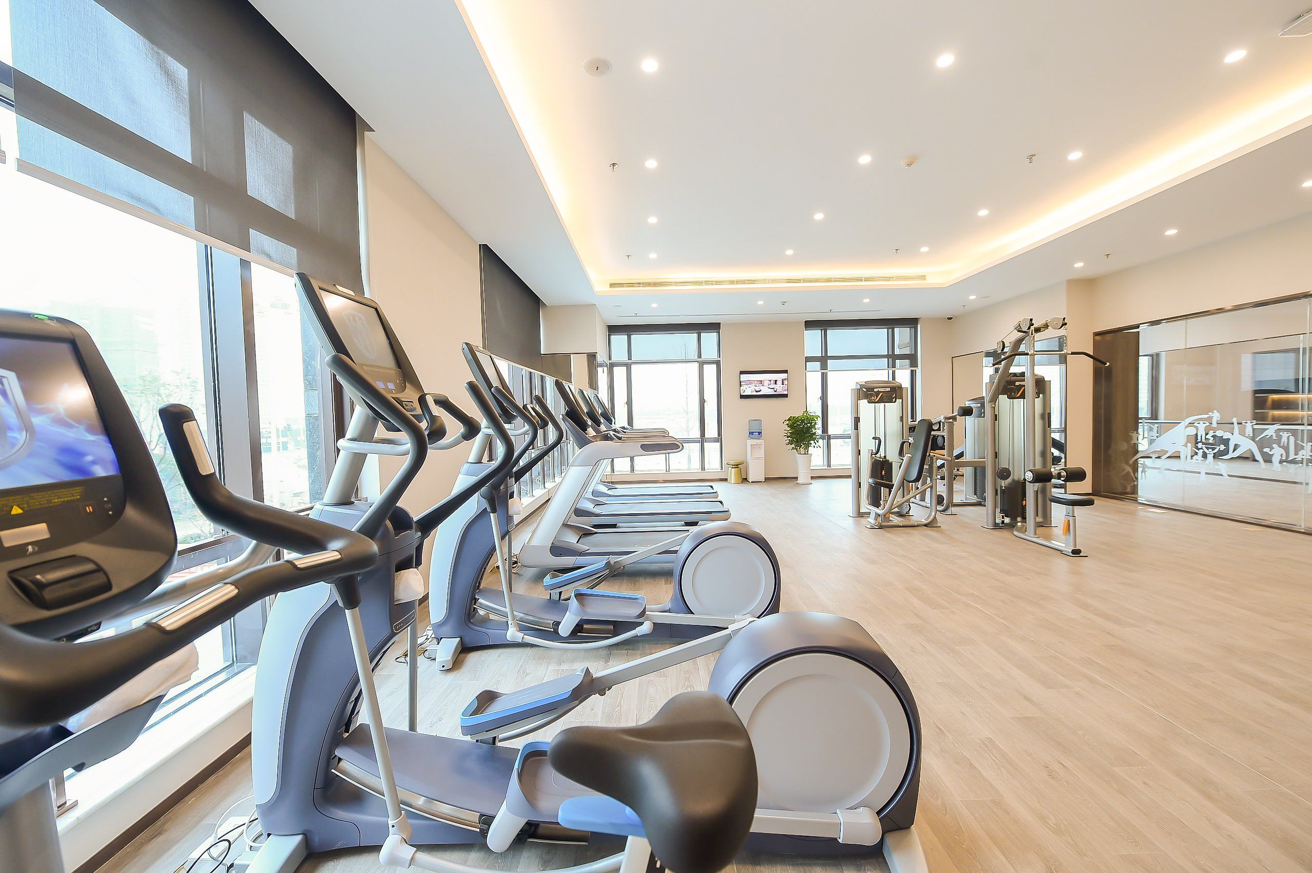 Un gimnasio con varias cintas de correr, máquinas de ejercicio y un amplio ventanal con espejos.