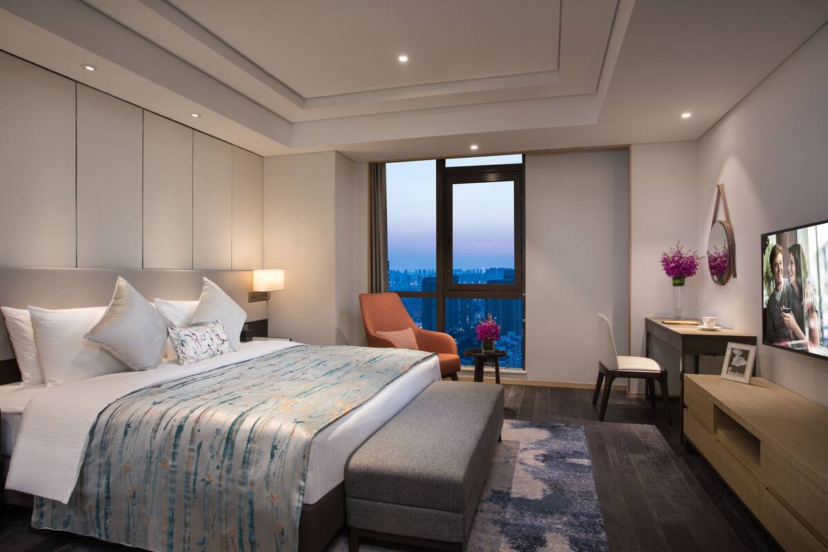 A modern hotel room with a large bed, a seating area with a chair and a small table, a TV, and a view of the city through a large window.
