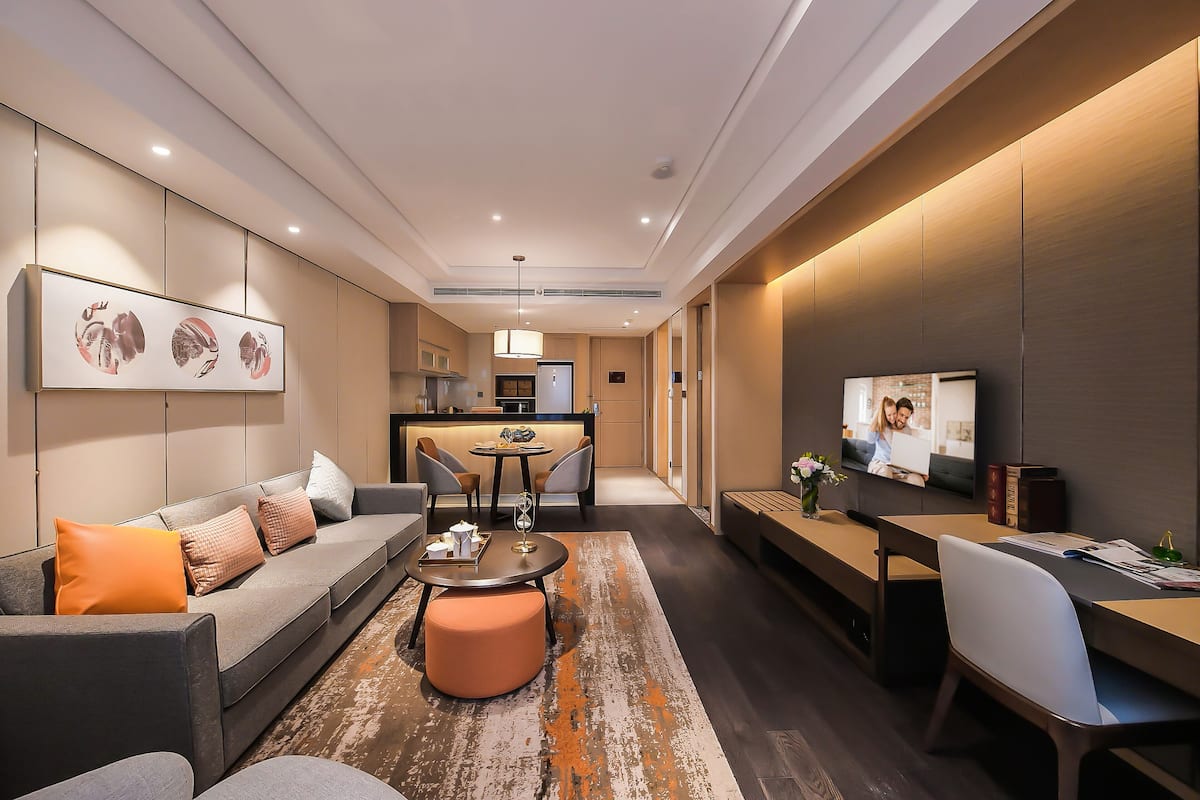A modern hotel room with a sofa, ottoman, dining table, and a TV.