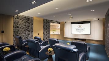 Una habitación de hotel moderna con una pantalla grande que muestra el nombre del hotel, una zona de estar con sillones de cuero y una mesita con flores.