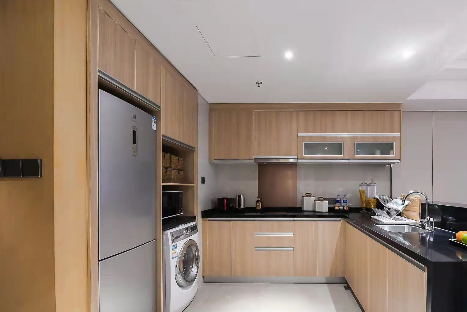 A modern kitchen with wooden cabinets, a stainless steel refrigerator, a built-in washing machine, and a black countertop.