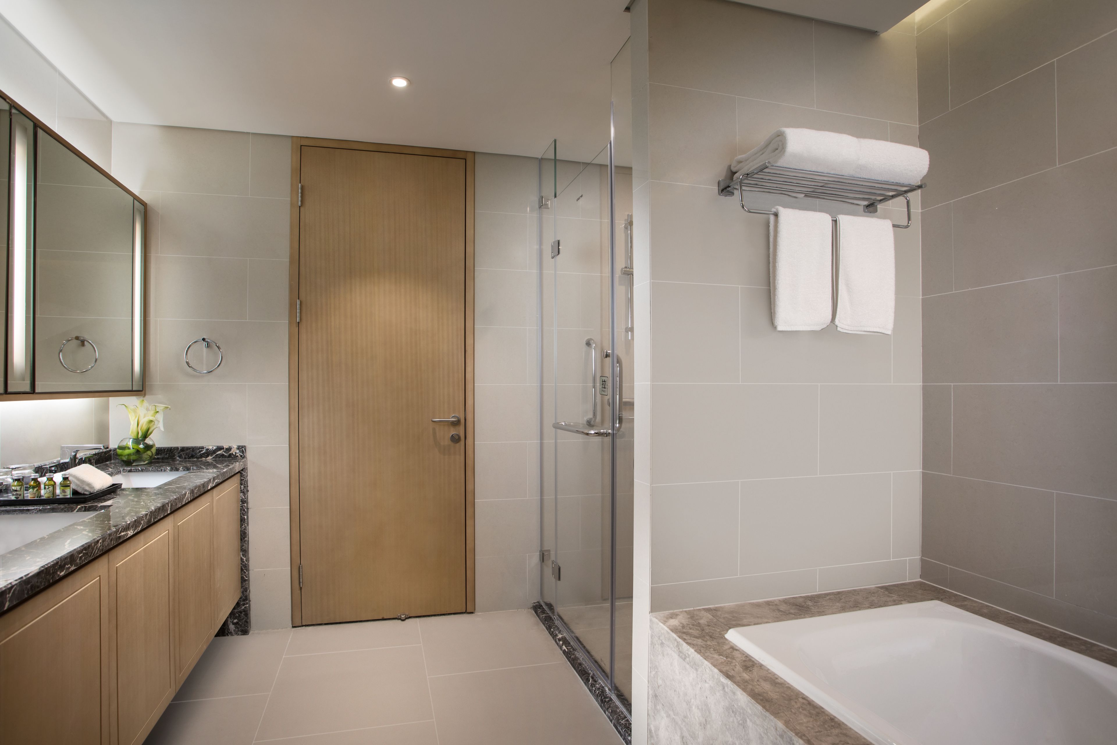 A modern bathroom with a glass-enclosed shower, a wooden door, a marble countertop, and a white bathtub.