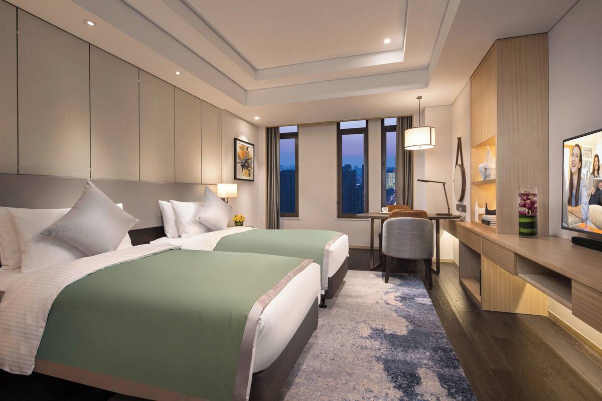 A hotel room with two beds, a desk, a chair, a TV, and a large window with a city view.
