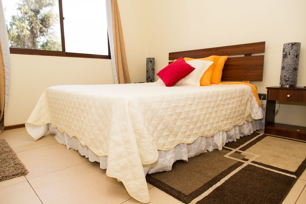 A neatly made bed with a white quilt, a wooden headboard, and a brown and beige rug on a tiled floor.
