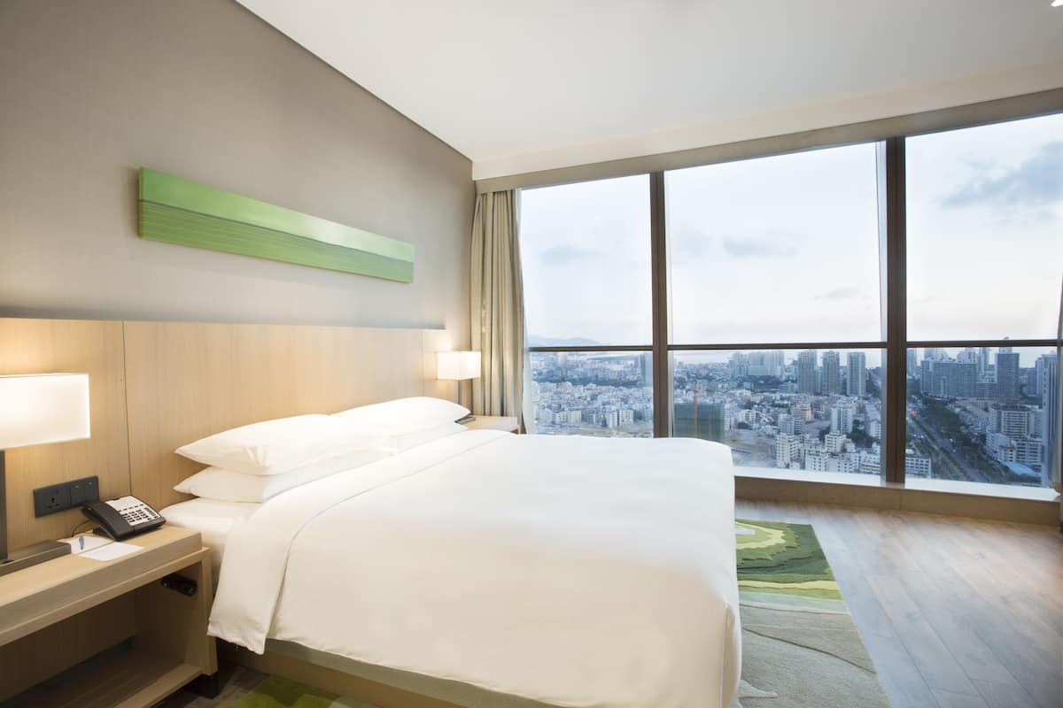 Hotelkamer met een groot bed, nachtkastje met telefoon en uitzicht op een stadsgezicht door een groot raam.