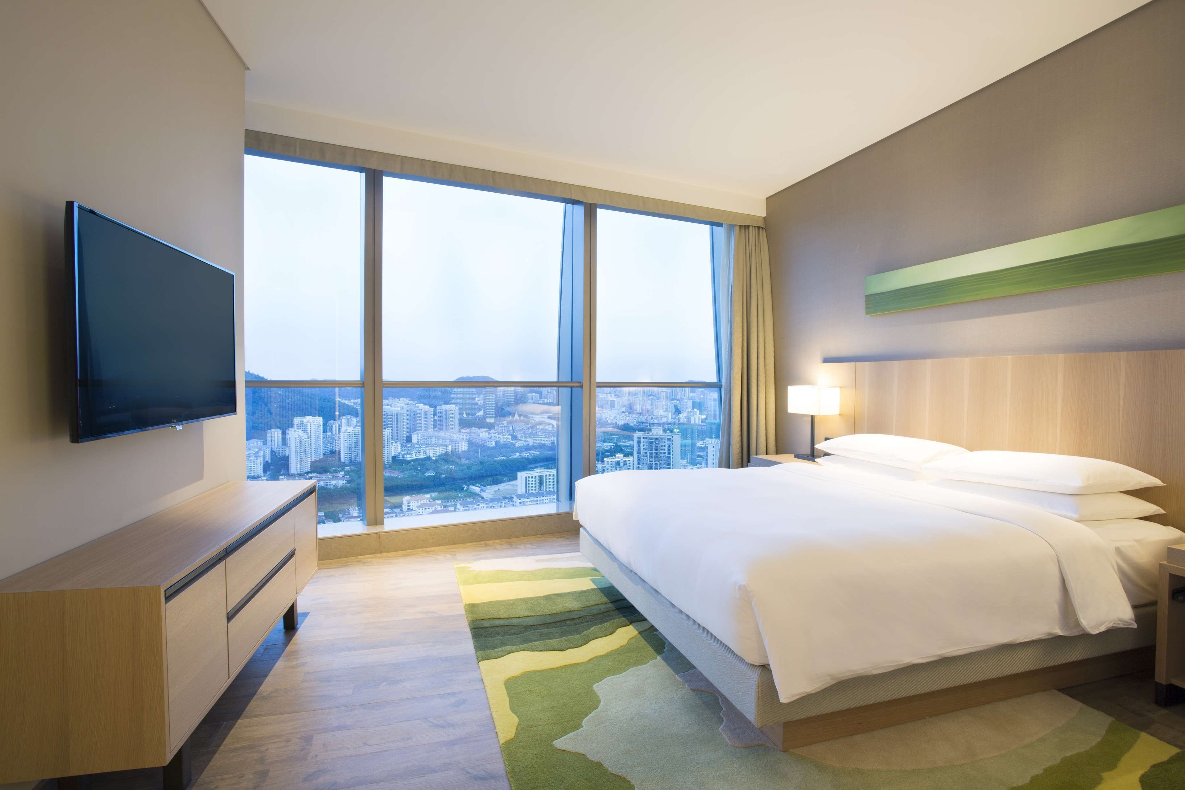 A hotel room with a large window, a bed, a TV, a nightstand, and a wooden dresser.