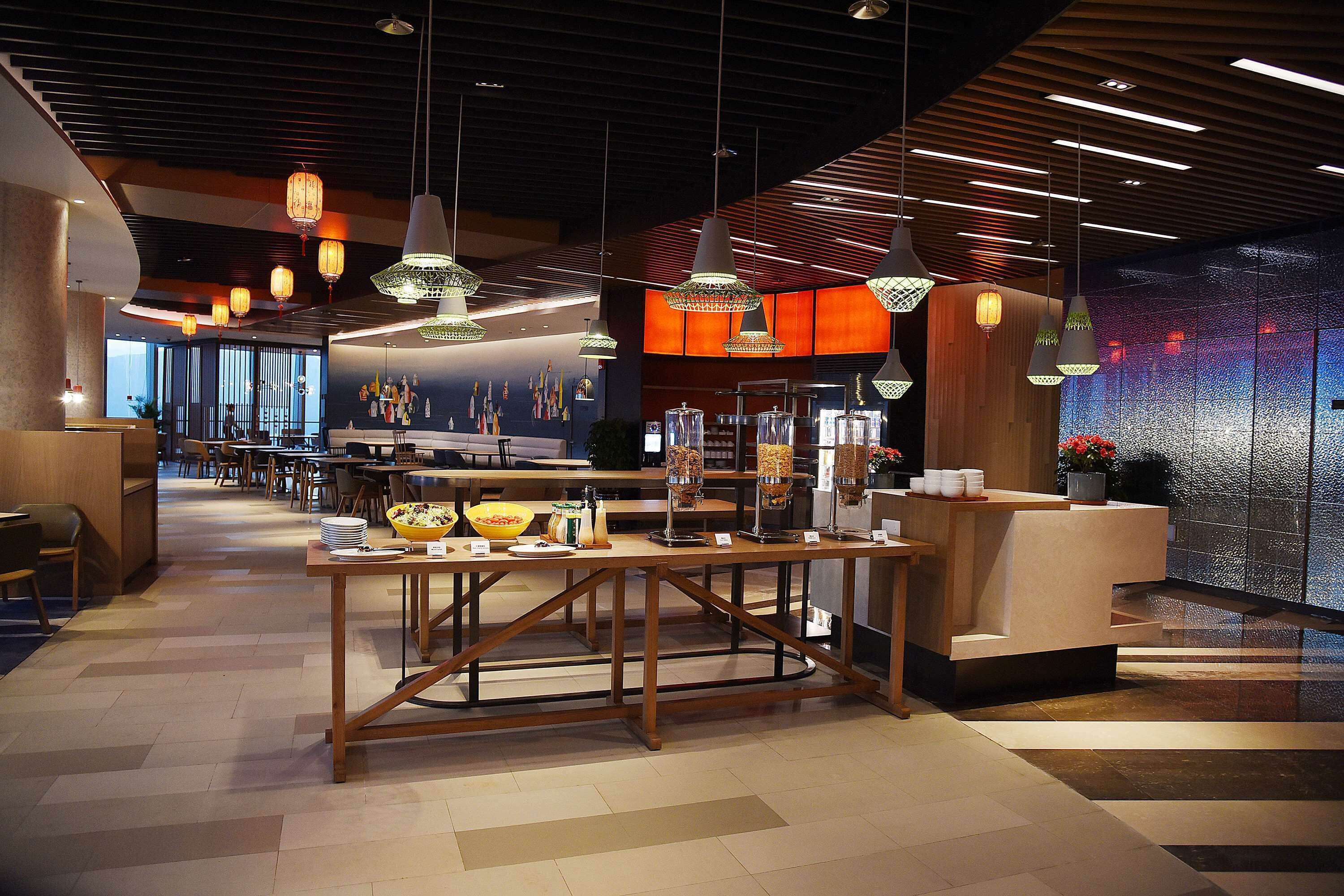 A modern hotel lobby with a reception desk, dining area, and a large window offering a view of the cityscape.